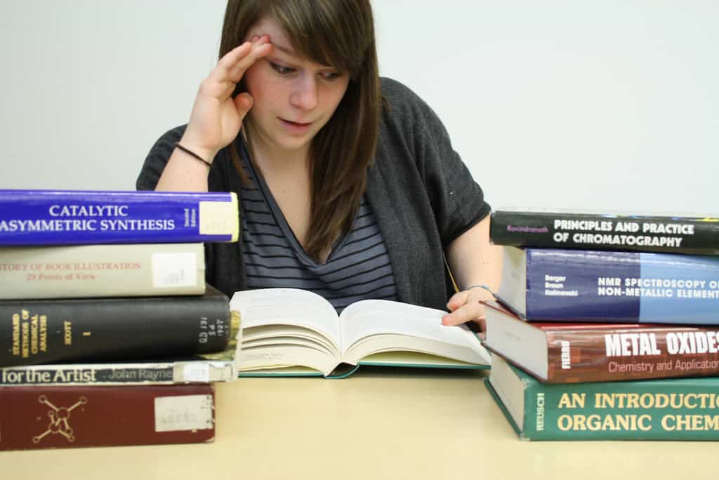 Female student looking skeptically at textbook, while other textbooks surround her - The idiot