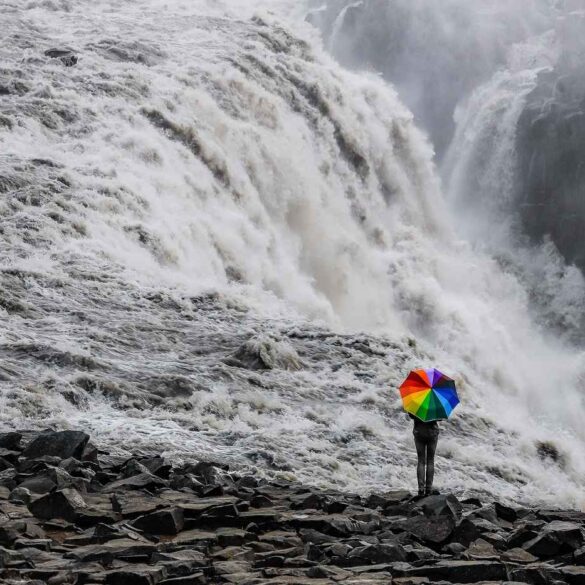 Person walking with rainbow-coloured umbrella near waterfall - Some Simple Truths About Mindfully Being Who We Are