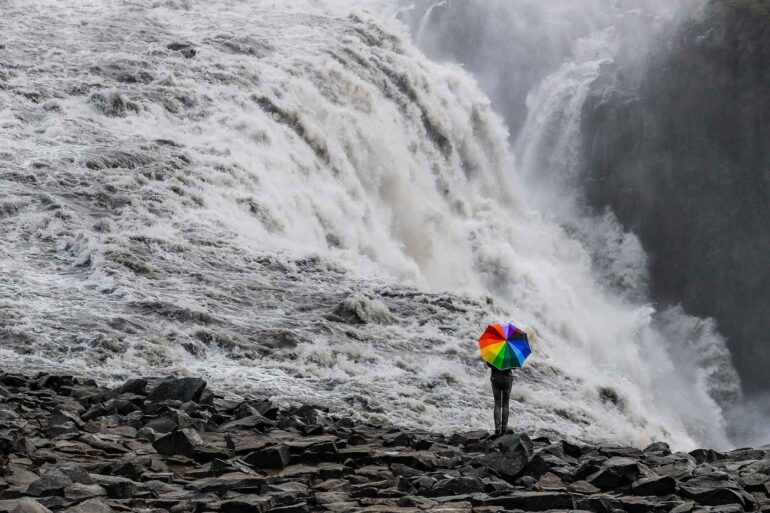 Person walking with rainbow-coloured umbrella near waterfall - Some Simple Truths About Mindfully Being Who We Are
