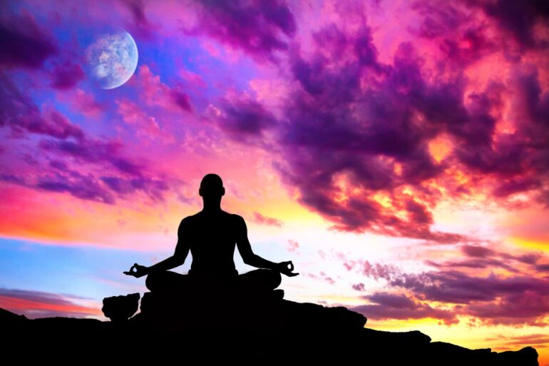 Silhouette meditating on rocks below swirly purple sky - 8 Mindful Practices To Help You Manifest the Life You Desire