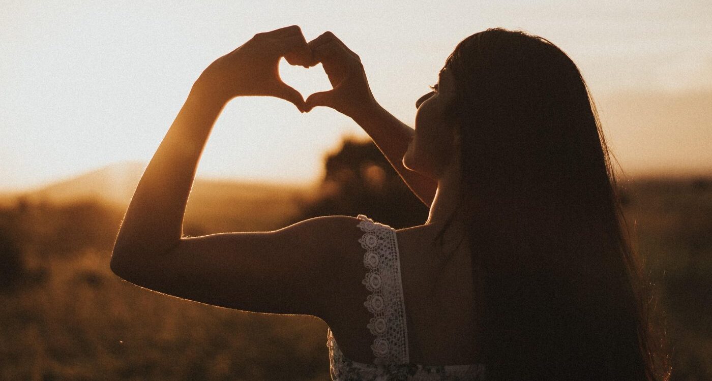 Woman standing alone outdoors making heart shape with hands - An Invitation to Women: Let's Be Gentle With Ourselves Now