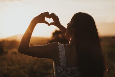 Woman standing alone outdoors making heart shape with hands - An Invitation to Women: Let's Be Gentle With Ourselves Now